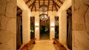 An interior with stone walls, wooden benches, a thatched ceiling, and a central light fixture.