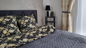 A bed with a black and yellow floral comforter, a grey quilted bedspread, a black lamp on a small table beside the bed, and a curtain on the window. - A comfortable apartment close to the seaside beach (Gaski)