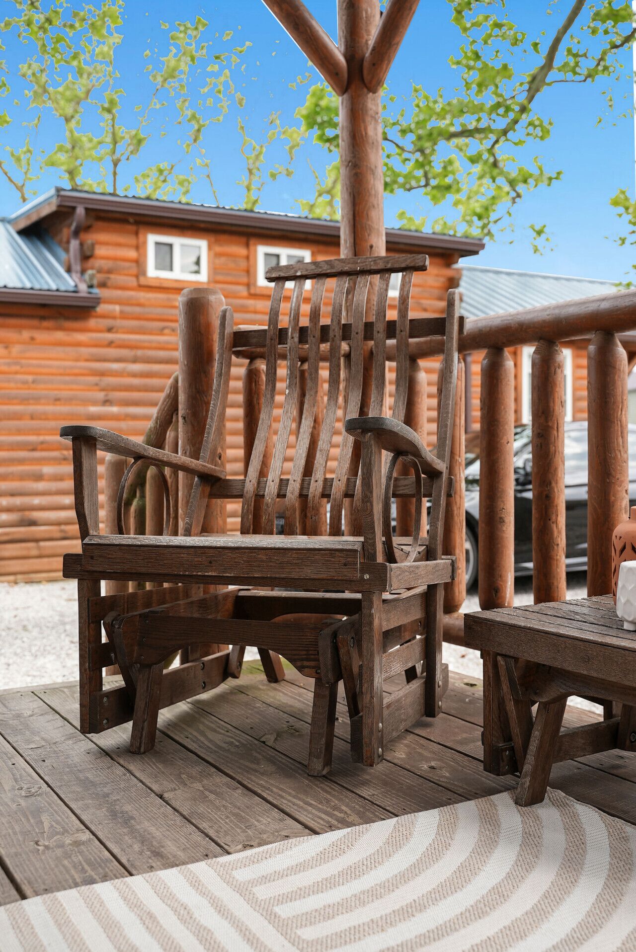A wooden Adirondack chair and a small table on a wooden deck with a striped rug, in front of a log cabin with a metal roof.