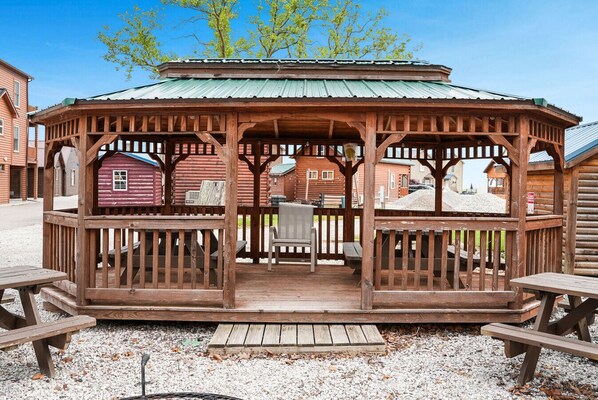 Un kiosque en bois surmonté d’un toit vert, entouré d’autres bâtiments en bois et agrémenté de bancs et d’une table.
