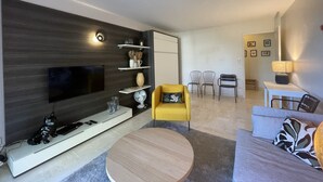 Un salon moderne avec un téléviseur à écran plat, un fauteuil jaune, une table basse ronde et un canapé. On y trouve des étagères ornées d’objets décoratifs et, au fond, un coin repas.