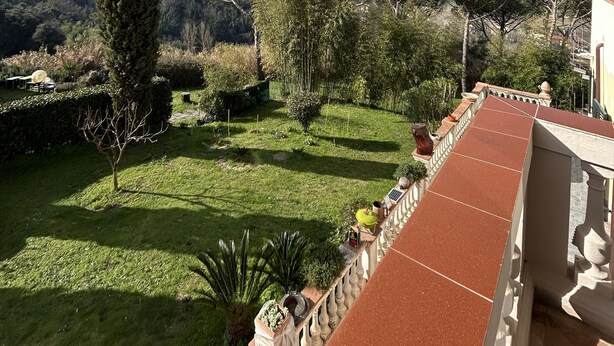 Un balcon doté d’un sol carrelé de tuiles rouges, donnant sur un jardin bien entretenu où poussent différentes plantes et s’élève un grand arbre.