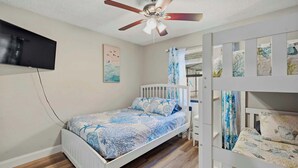 Un dormitorio con una cama, un ventilador de techo, un televisor montado en la pared y una cuna con una colcha estampada.