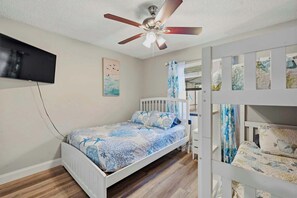 Un dormitorio con una cama, un ventilador de techo, un televisor montado en la pared y una cuna con una colcha estampada.