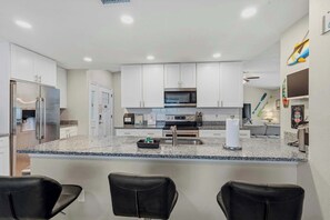 A modern kitchen with white cabinets, a stainless steel refrigerator, a microwave, and a gas stove. There are three black leather chairs in the foreground.