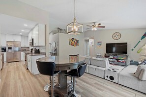 A modern kitchen with a stainless steel refrigerator, a dining area with a round table and bar stools, a living room with a large sofa and a flat-screen TV, and a ceiling fan.