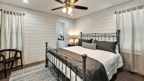 Un dormitorio con cama, mesita de noche, silla, ventilador de techo y ventana con cortinas.