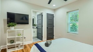 A bedroom with a bed, a ceiling fan, a TV, a window with blinds, and a door leading to a bathroom. - Elegant Atlanta Haven | 3 Bedroom Near Downtown (Atlanta)