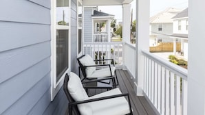 Une véranda couverte avec deux fauteuils en osier noir et des coussins blancs, des balustrades blanches et une vue sur les maisons voisines.