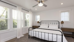 Une chambre à coucher comprenant un lit, deux tables de chevet, un ventilateur de plafond et de grandes fenêtres avec des rideaux blancs.