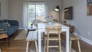 A modern living room with a white table, wooden chairs, a blue sofa, and a large window with curtains.