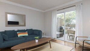 A living room with a dark green sofa, a wooden coffee table, and a small plant. There is a sliding glass door leading to an outdoor patio area with a table and chairs.