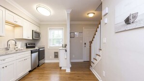 A kitchen with white cabinets, a microwave, a sink, and a dishwasher. There is a window with blinds, a door with a picture frame, and a staircase with wooden steps.