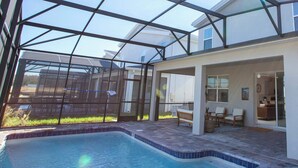 Una piscina con cerramiento de vidrio, un área de patio con mobiliario y vista a un edificio residencial al fondo.