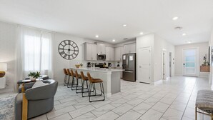 Una cocina moderna con armarios blancos, una isla central con taburetes, un reloj de pared grande y una zona de comedor con sofá y sillas.