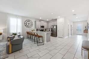 A modern kitchen with white cabinets, a central island with bar stools, a large wall clock, and a dining area with a sofa and chairs.