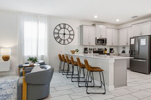 A modern kitchen with white cabinets, a central island with bar stools, a large wall clock, and a microwave.