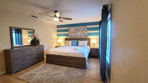 A bedroom with a bed, dresser, ceiling fan, and artwork on the wall.