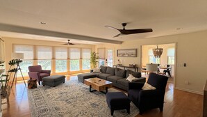 A spacious living room with a sectional sofa, a coffee table, and a large rug. There are ceiling fans, a dining area, and large windows with blinds.