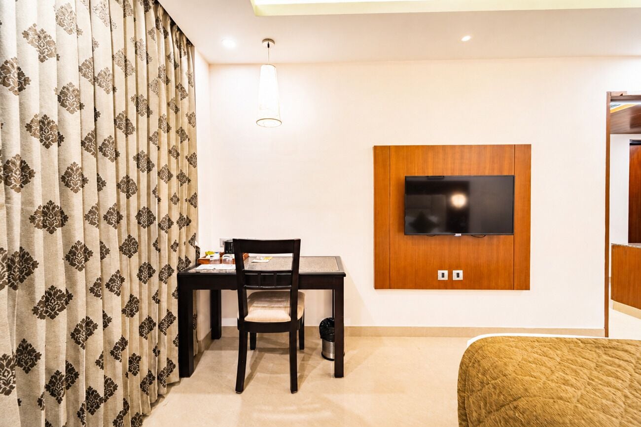 A hotel room with a bed, a TV mounted on the wall, a desk with a chair, and a curtain with a pattern.