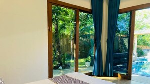 Ein Schlafzimmer mit einem großen Bett, Blick auf einen tropischen Garten durch Glastüren und ein Pool im Hintergrund.
