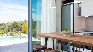 A modern kitchen with a wooden table and bar stools, a bowl of fruit, and a view of a swimming pool and greenery outside.