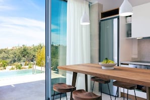 A modern kitchen with a wooden table and bar stools, a bowl of fruit, and a view of a swimming pool and greenery outside.