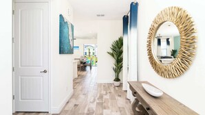 Een hal met een witte deur, een blauw abstract schilderij, een potplant, een houten bank en een spiegel met zonneschijnbelichting.