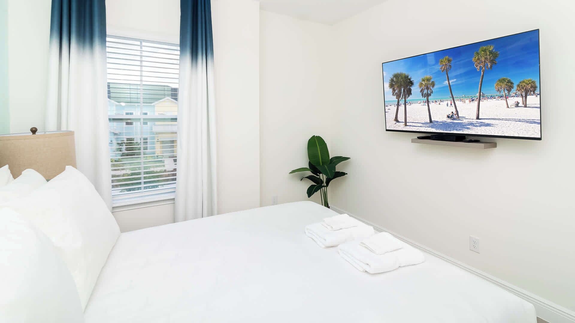 A hotel room with a bed, a TV on the wall, a window with curtains, and a plant.