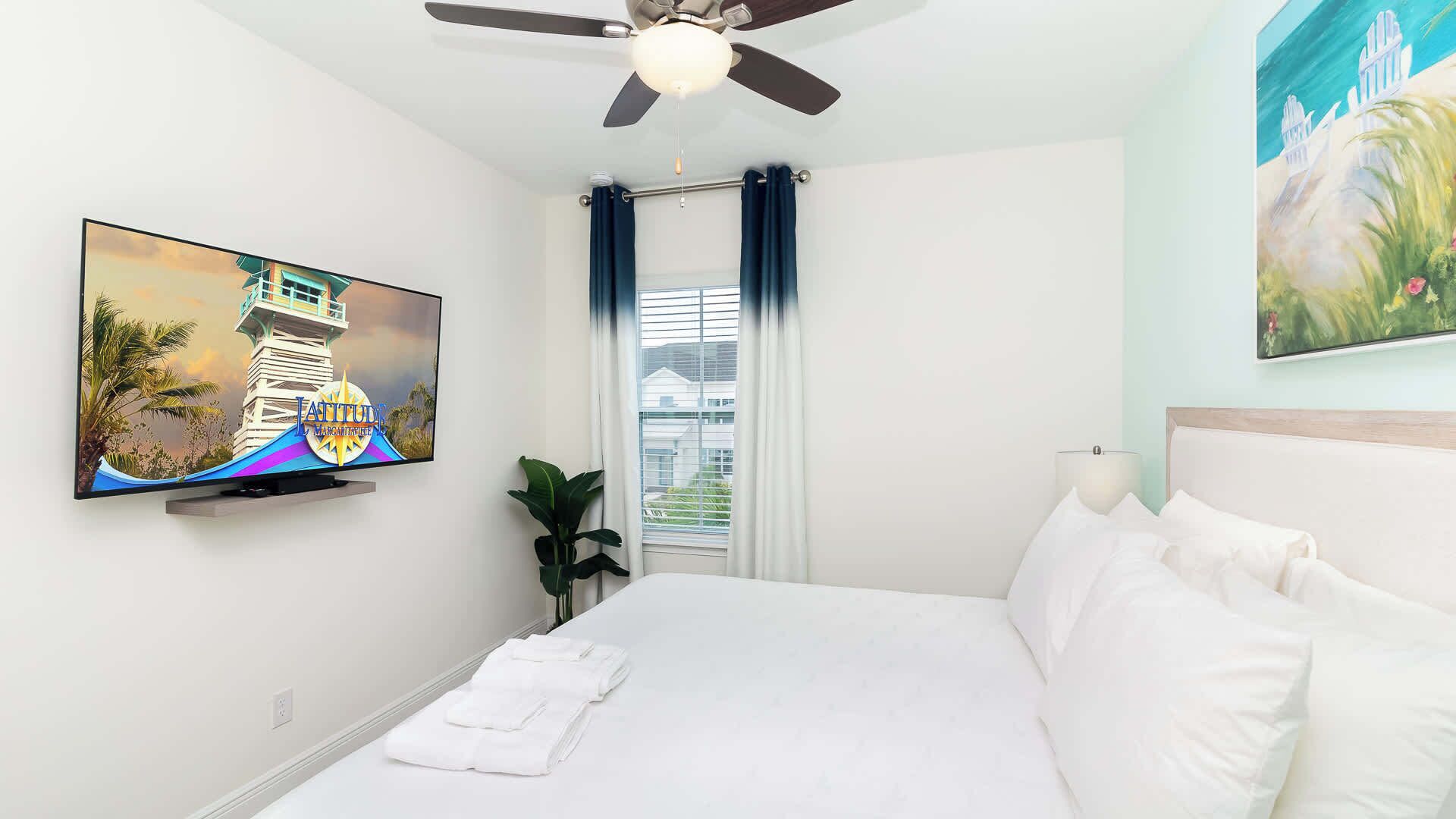 A hotel room with a bed, a ceiling fan, a TV on the wall, a window with curtains, and a painting on the wall.