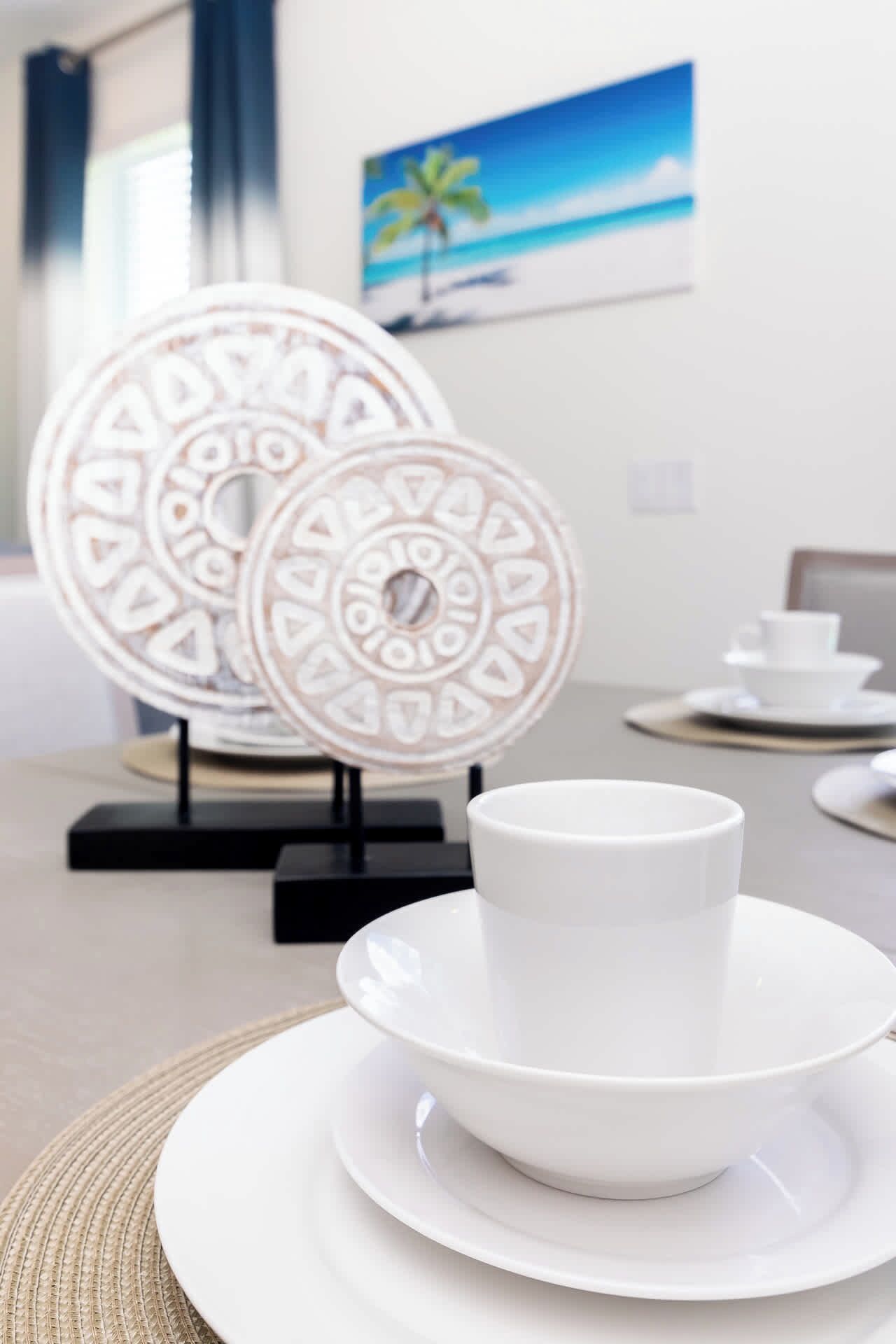 Un coin repas avec de l’assiette blanche et une tasse sur une table, une décoration murale et une fenêtre avec des rideaux.