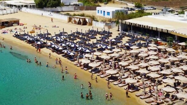 Una spiaggia con numerosi lettini e ombrelloni, una riva sabbiosa e alcuni edifici sullo sfondo.