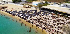 A beach with numerous sun loungers and umbrellas, a sandy shore, and a few buildings in the background. - 1Path houses Mykonos, Beach Cycladic home near the Super Paradise beach sand (Mykonos)