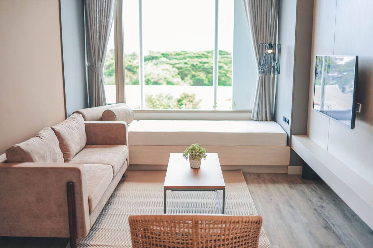 A modern living room with a sofa, a small table with a plant, a wicker chair, and a TV mounted on the wall.