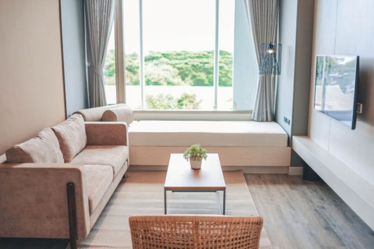 A modern living room with a sofa, a coffee table with a plant, and a view through a large window.