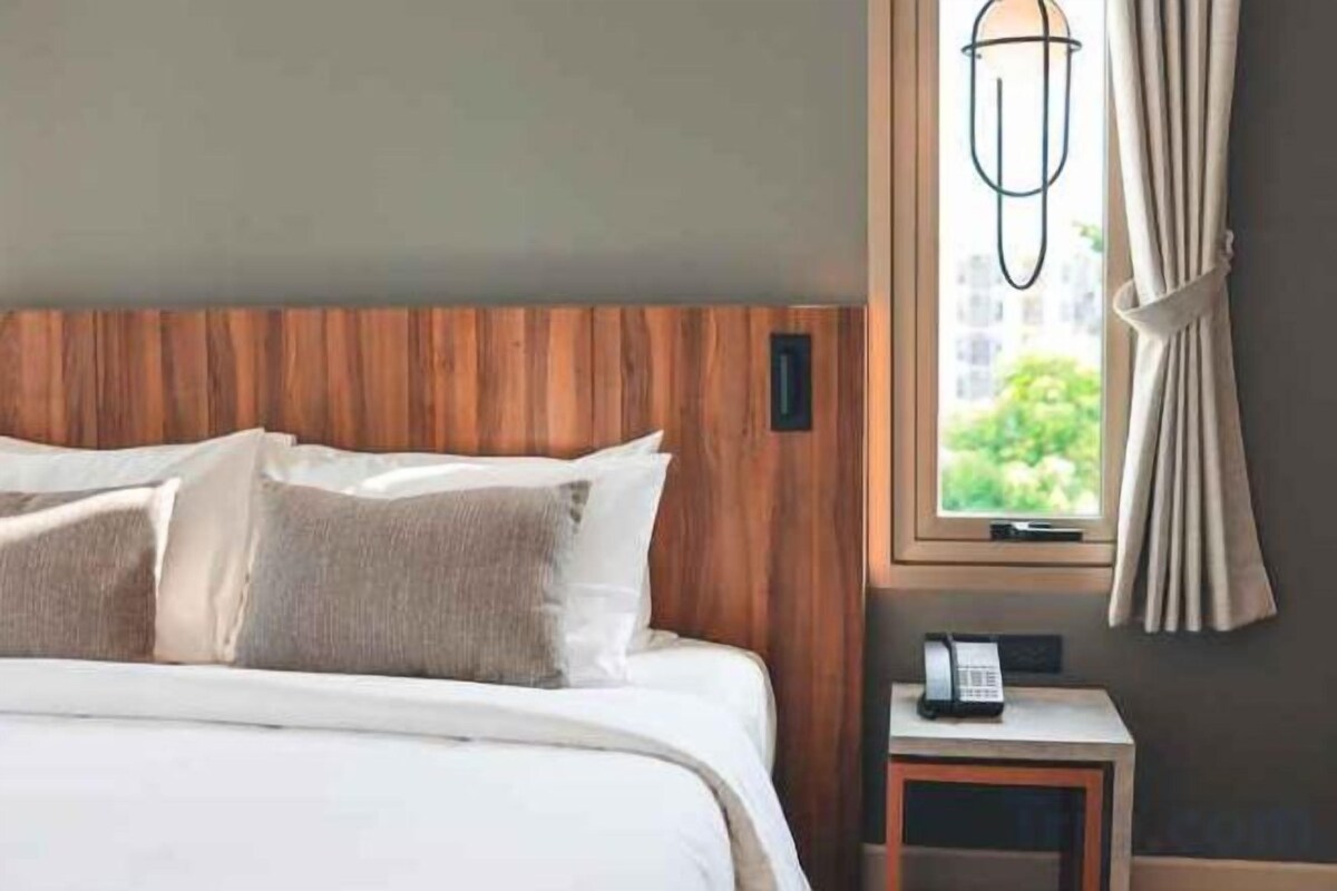 A hotel room with a wooden headboard, a bed with white bedding, a bedside table with a telephone, and a window with curtains.