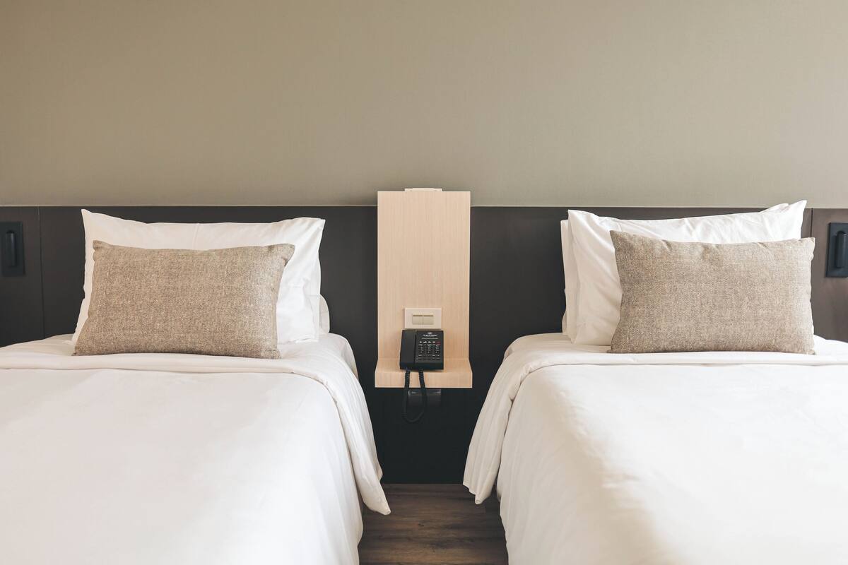 A hotel room with two beds, each with white bedding and a beige pillow. A wooden panel with a telephone is mounted on the wall between the beds.