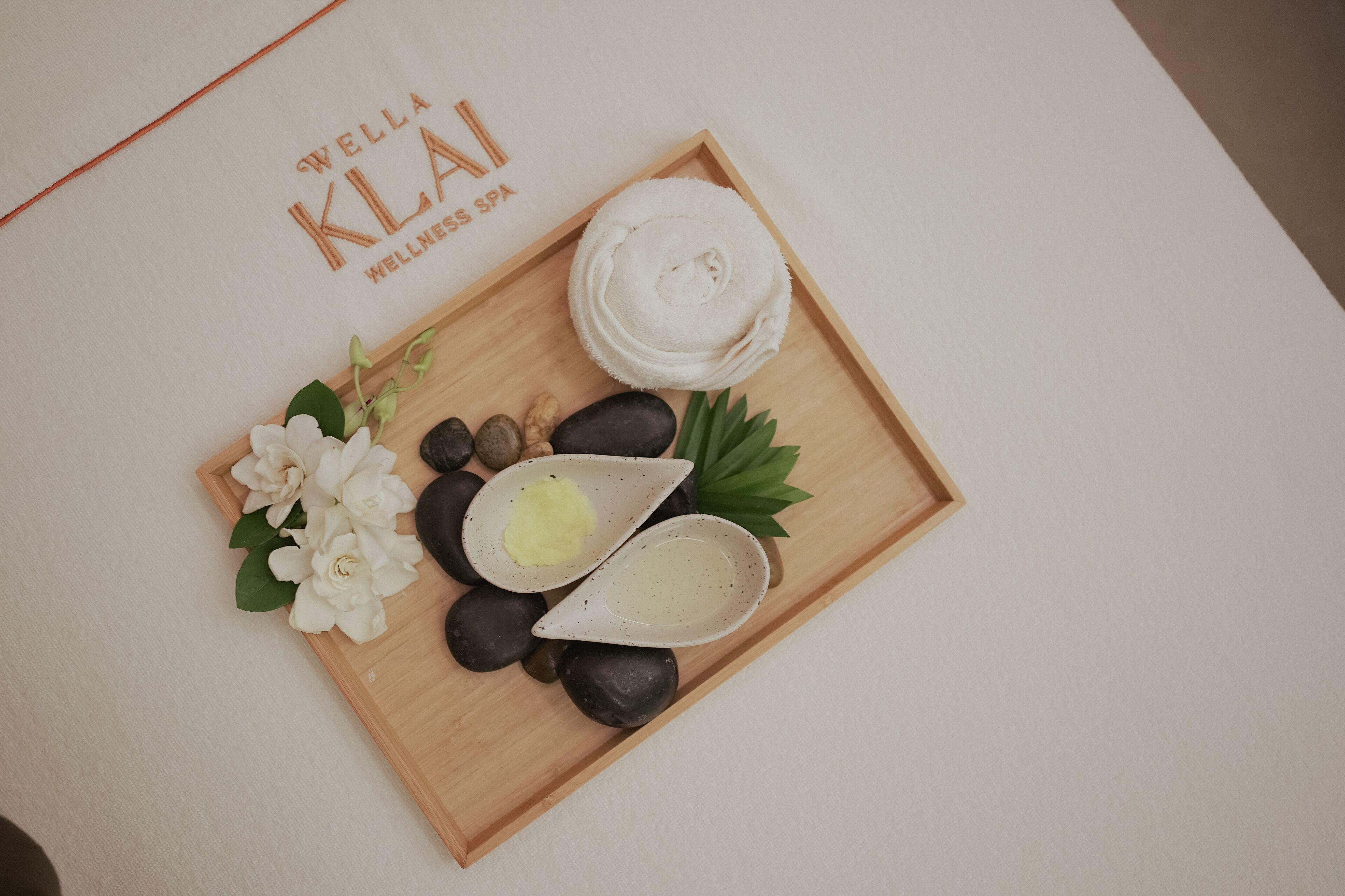 A wooden tray with a rolled towel, two small bowls, and white flowers on a white surface.