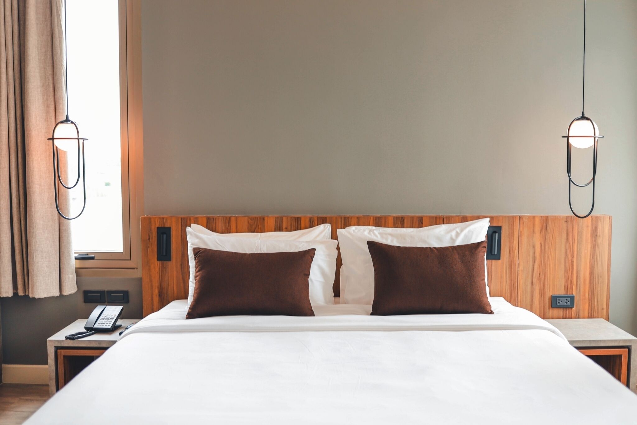 Quarto de hotel com cabeceira de madeira, duas almofadas, mesa de cabeceira com telefone e janela com cortinas.