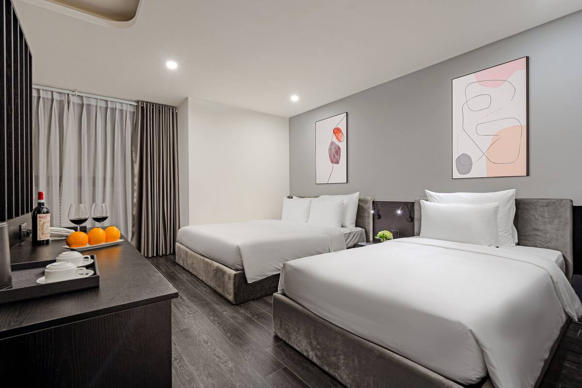 A hotel room with two beds, a desk with a bottle of wine and glasses, and a tray with cups and oranges.