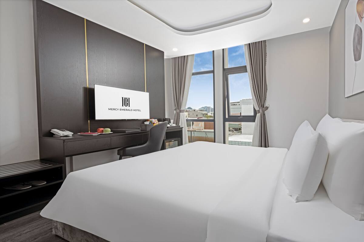 A hotel room with a large bed, a desk with a telephone, and a TV displaying the hotel name.