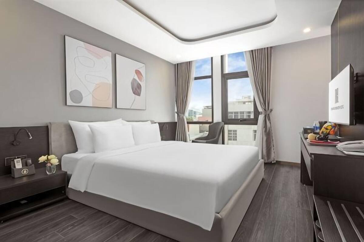 A modern hotel room with a large bed, a desk with a chair, a vanity, and a window with curtains.