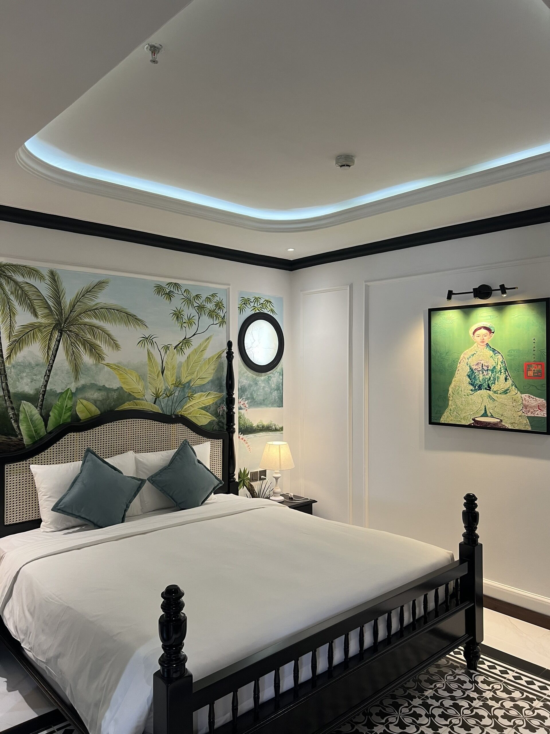 A neatly arranged bedroom with a bed, a nightstand, a wall-mounted lamp, a painting, and a ceiling light fixture.
