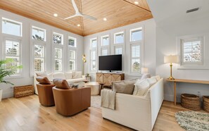 A spacious living room with a large flat-screen TV, multiple seating options including a sofa and two armchairs, a ceiling fan, and large windows providing natural light.