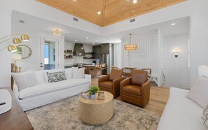 A spacious living area with a white sofa, brown leather chairs, and a round coffee table. A kitchen is visible in the background with modern appliances.