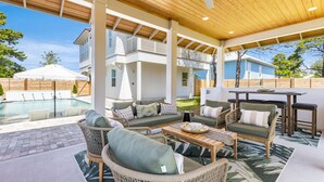Un espacio de estar al aire libre con techo, mobiliario de mimbre, piscina y vista a una casa al fondo.