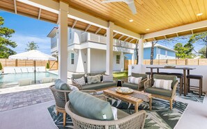 A covered outdoor seating area with wicker furniture, a pool, and a view of a house in the background.