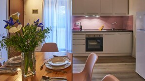 Ein Essbereich mit Holztisch, gedeckt für zwei Personen, einer Blumenvase und Blick in eine Küche mit weißen Schränken und einer rosa Spritzschutzleiste.