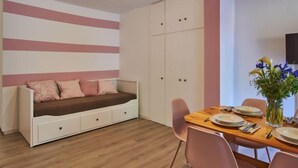 Ein Zimmer mit Holzboden, einer Couch mit Stauraum, einem Esstisch gedeckt mit Tellern und einer Blumenvase, sowie einem an der Wand montierten Fernseher.