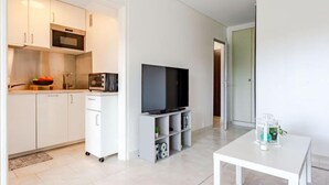 Een moderne keuken met witte kasten, een magnetron en een inbouwoven. Er staat een televisie op een dressoir met opbergplanken eronder. Een witte tafel bevindt zich op de voorgrond.